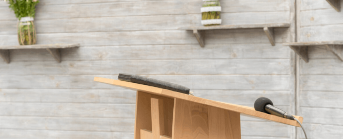 A wooden lectern with a microphone and book, set against a light timber wall with floral arrangements.